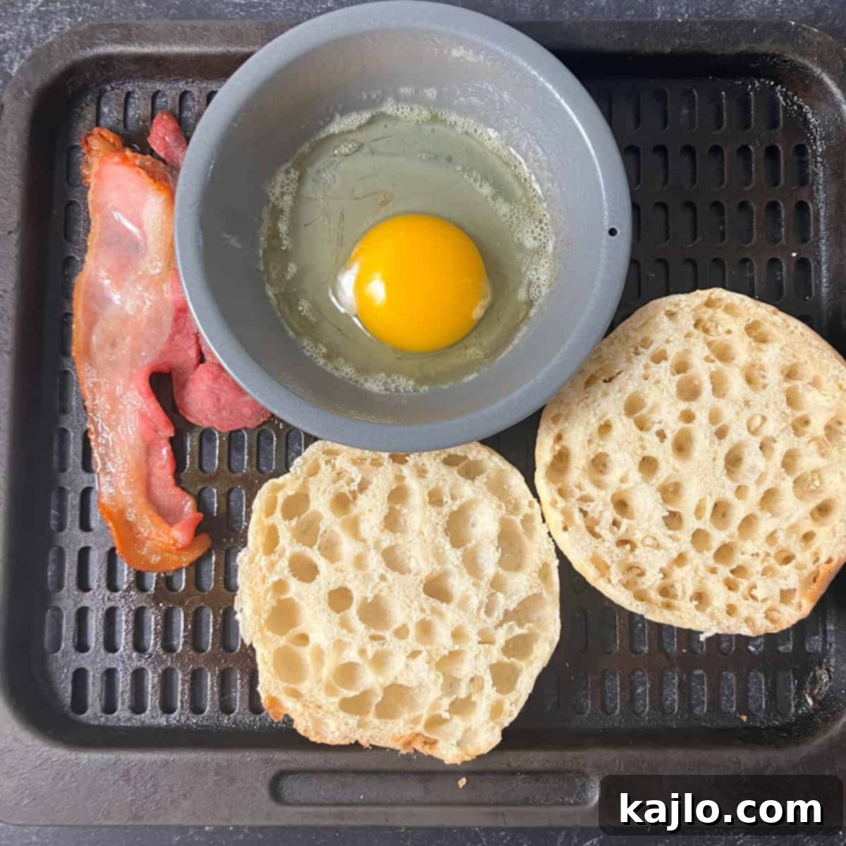 Egg and English muffin cooking in the air fryer alongside partially cooked bacon.